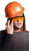 A woman in an orange helmet and protective eyeglasses is talking and holding a pen and folder in her hands on a white background. Features of work of the engineer Vertical video