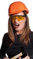 A beautiful woman in orange eyeglasses and helmet is holding a hammer in her hands and hitting herself by it in her hand on a white background in the studio. Engineer Vertical video