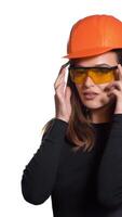 A joyful woman in orange helmet for building is trying on goggles and posing in it on a white background in the studio. Vertical video