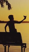 a little boy is sitting on the bench and playing with a paper boat on the background of evening landscape by the lake in the summer. Sunset Vertical video