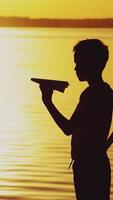 A little boy is holding a paper airplane in his hand and playing with him on the bank of the river at sunset on the background of the solar track. Vertical video