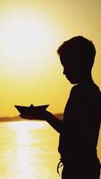 A little boy is holding an origami in the form of a paper boat in his hands and raising up it to the level of the sun on the background of the lake. Silhouette of a guy at sunset Vertical video