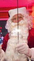 Santa indoors on New year tree background. Portrait of a real Santa Claus with white beard holding sparkling lights and looking at camera. Happy New Year. Vertical video