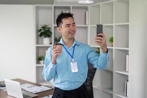 Cheerful Asian businessman taking a selfie while holding a coffee cup in a modern office environment. photo