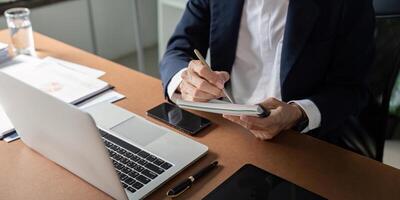 Elderly businessman taking notes in a notebook while working on a laptop at a modern office desk. photo