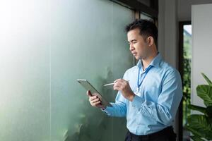 Focused Asian businessman using a tablet for analysis, standing by a frosted glass window in a modern office environment. photo