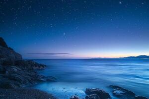 The sky transitions to twilight as stars emerge above a serene ocean. Gentle waves lap against the rocks, creating a peaceful atmosphere at dusk photo