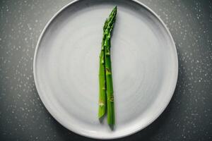 Bright green asparagus is arranged neatly on a plain white plate, showcasing its vibrant color against a textured, softly lit backdrop, emphasizing freshness photo