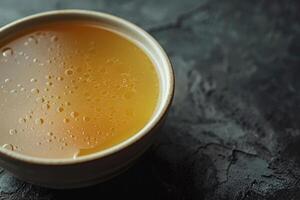 A simple bowl filled with warm golden broth sits on a dark textured surface. The broth displays tiny bubbles, suggesting warmth and comfort, perfect for a chilly day photo