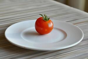 A vibrant red tomato is placed on a simple white plate, resting on a lightly textured wooden table, illuminated by natural light photo