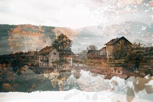 The tranquil countryside showcases charming houses surrounded by vibrant autumn foliage, mirrored beautifully in the still water below under a soft afternoon light photo