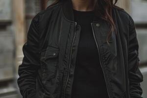 The individual is dressed in a fitted black bomber jacket paired with a black sweater, posing against an urban backdrop of subtle textures under an overcast sky photo