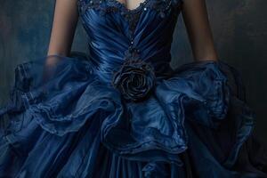 A person is wearing a stunning blue dress adorned with delicate embellishments, showcasing layers of flowing fabric and a rose accent. Soft lighting enhances the gown's elegance photo