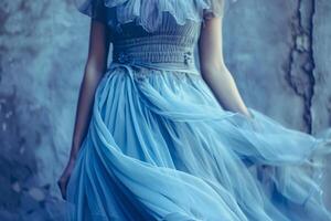 The woman stands elegantly, her delicate blue dress billowing gently around her. The soft fabric contrasts beautifully with the textured gray wall behind her, creating an enchanting mood photo