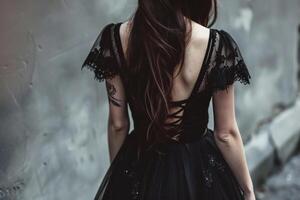 A woman wearing a black lace dress with delicate sleeves is positioned in front of a weathered gray wall. Her long hair cascades down her back, emphasizing the elegant design photo