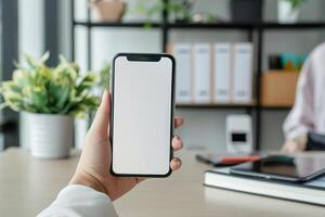 In a modern office environment, an individual displays a blank smartphone in one hand while surrounded by greenery and organized shelves, indicating a professional atmosphere photo