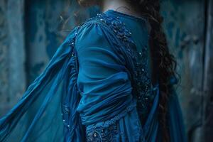 A figure adorned in an intricate blue dress stands against a weathered wall, the flowing fabric gracefully catching the light, hinting at a moment of serene reflection photo