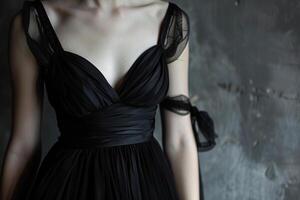A woman in an elegant black dress with delicate sheer straps stands against a textured gray backdrop, emphasizing the garment's flowing design and fabric details photo