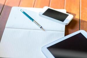 Workspace setup with a pen, notebook, and digital devices on a wooden table during bright daylight hours photo