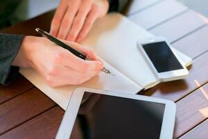 Individual writing notes with a pen on a notebook, surrounded by digital devices on a wooden table photo