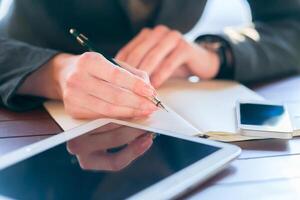 Person writing notes with a pen while seated at a desk with a tablet and smartphone nearby in a well-lit space photo