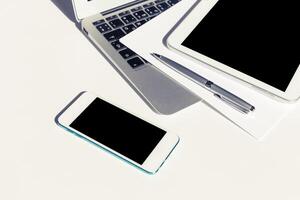 Workspace with electronic devices including smartphone, tablet, and laptop on a white surface during daylight hours photo