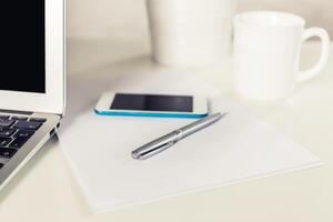 Workspace setup with laptop, smartphone, pen, and empty paper on a table in a well-lit room photo