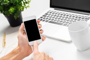 Person using smartphone while working at desk with laptop and plant in modern office setting photo