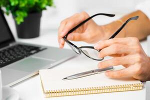 Man adjusting glasses while preparing to write notes at a laptop on a desk with a small plant photo