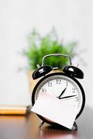 Vintage black alarm clock sitting on a wooden table with a note and greenery in the background during daylight photo