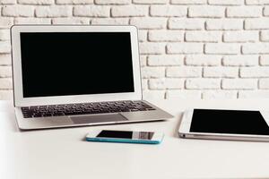 Workspace featuring a laptop, smartphone, and tablet on a white brick background during the afternoon photo