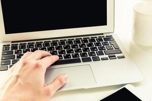 Hand typing on a laptop keyboard while sitting at a desk with a cup nearby in a bright workspace photo