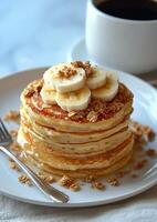 Stack of fluffy pancakes topped with banana slices and granola beside a cup of coffee photo