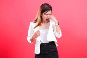 A woman wearing a white jacket and black pants is holding her nose and sticking out her tongue, isolated on a red background photo