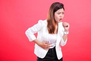 A woman wearing a white jacket coughs while holding her stomach. The image is of a person coughing with discomfort, isolated on a red background. photo