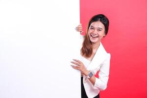A smiling woman peeking behind a large blank white sign, she is wearing a white blazer and a smart watch and her nails are metallic, isolated on a red background photo