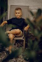 A boy in a black T-shirt sits in a garden chair in the backyard of a house photo