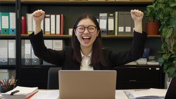 A asian young woman sitting at a desk in an office with her arms raised in the air. She is wearing a black blazer and a white shirt, and has a big smile on her face. photo