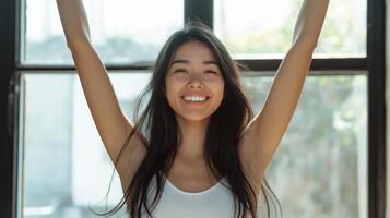 portrait of a asia young woman with long dark hair, standing in front of a large window with her arms raised above her head photo