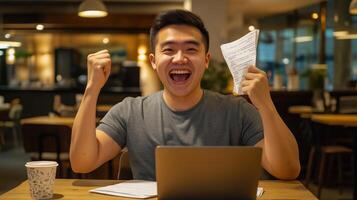 asian man sitting at a table in a cafe or restaurant. He is wearing a grey t-shirt and has a laptop in front of him. photo