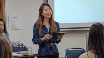 asian young woman standing in front of a group of people in a classroom. She is holding a tablet and appears photo