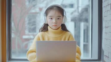 asian young girl sitting on a windowsill with a laptop in front of her. She is wearing a yellow sweater and has a pair of white headphones on her head photo