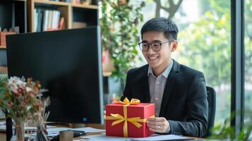 asian young man in a suit and glasses sitting at a desk in front of a computer monitor. He is holding a red gift box with a yellow ribbon photo