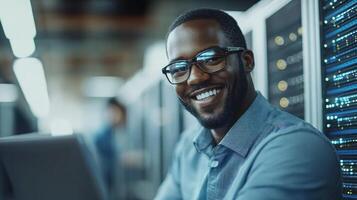 Technician with glasses smiles while seated near servers, providing support to clients in a data center photo