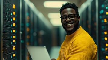 Man in a yellow sweater smiles while working on a laptop in a busy server room filled with equipment photo