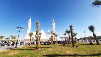 Madinah, Saudi Arabia on 28 February 2024. Quba Mosque. White Mosque under Blue Sky Palm Trees and People Gathering photo
