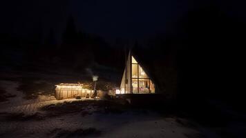un cuadro cabina brillante a noche con grande iluminado ventanas, rodeado por oscuridad y nieve. un pequeño de madera estructura emite fumar, agregando calor y encanto a el pacífico invierno escena video