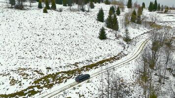 Car driving on snowy rural road, captured from aerial view, surrounded by sparse trees and hills. The black car moves along a winding snow-covered path with patches of green grass visible video