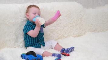 little boy sitting on the sofa drinking milk from a bottle and playing with a toy video