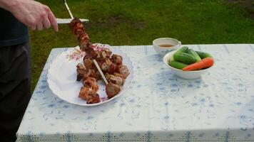 Cook removing pieces of shashlik, ready to eat grilled meat, from a metal skewer onto a white plate on a table with cucumbers and carrots, outdoors video